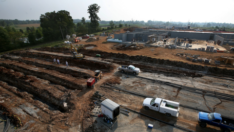 aerial photo in which construction crew digs long trenches that run parallel to a large uncompleted building.