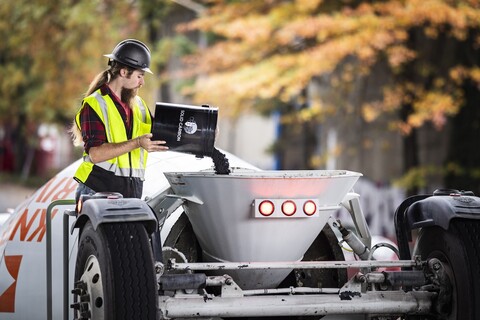 A worker pouring a bucket of black powder into a larger concrete mixer.