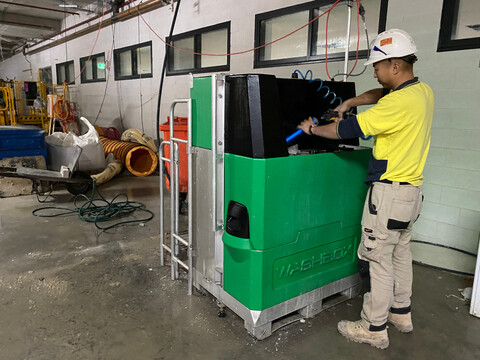 A construction worker dressed in a yellow top washes tools at arm level in a large green plastic box resting on the floor.
