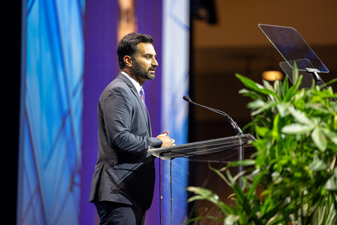 a man in a gray suit stands in at a podium. a large plant is on the right, and a luminous blue backdrop is behind him.